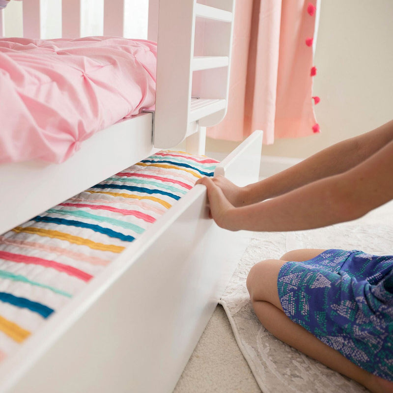 Child pulling out storage drawer under white bed with colorful linens in kids' bedroom.