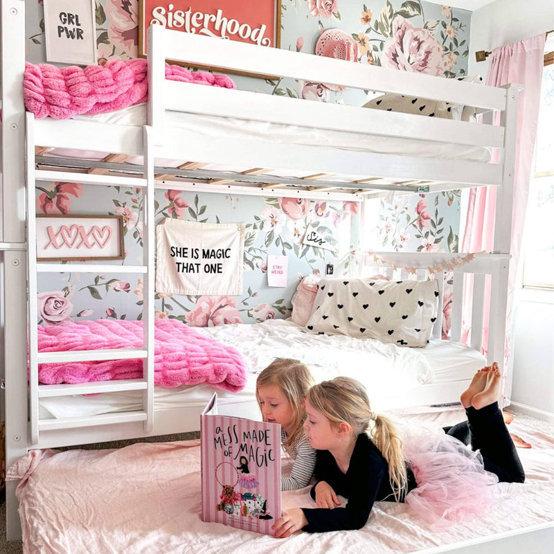 Two young girls reading a book in a floral-themed bedroom with a white bunk bed and pink decor.