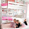 Two girls reading on a white bunk bed in a pink floral bedroom with plush pink blankets, heart-print pillow and girly kids room decor ideas