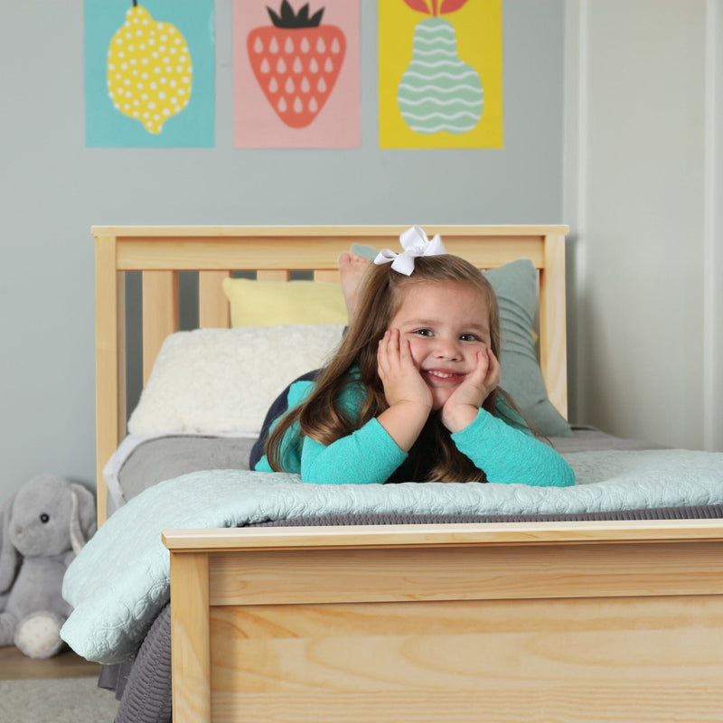Smiling young girl lying on a wooden children's bed with mint bedding, plush elephant toy, and colorful fruit wall art, cozy kids' bedroom and nursery decor