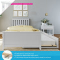 White children's wooden bed with pull-out trundle storage, yellow polka-dot bedding, solid pine frame and countersunk color-matched hardware beside a white nightstand with vase and books against a grey wall with botanical prints and gold pendant light