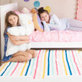 Two smiling girls lounging on a pink bunk bed with colorful striped bedding and plush pillows in a bright girls' bedroom — kids bedding, bunk bed, bedroom decor, sleepover setup