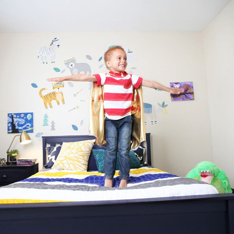 Child in a red and white striped shirt with a golden cape jumping on a bed in a colorful bedroom with animal wall decals.