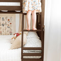 Child standing barefoot on wooden bunk bed ladder in cozy kids bedroom with patterned bedding and botanical wall art