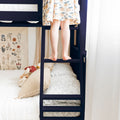 Child standing on navy bunk bed ladder in bright kids' bedroom, wearing a patterned dress with botanical wall art and cozy pillows