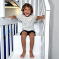 Happy young child with curly hair sitting on white bunk bed stairs in bright kids bedroom, smiling and holding the railing