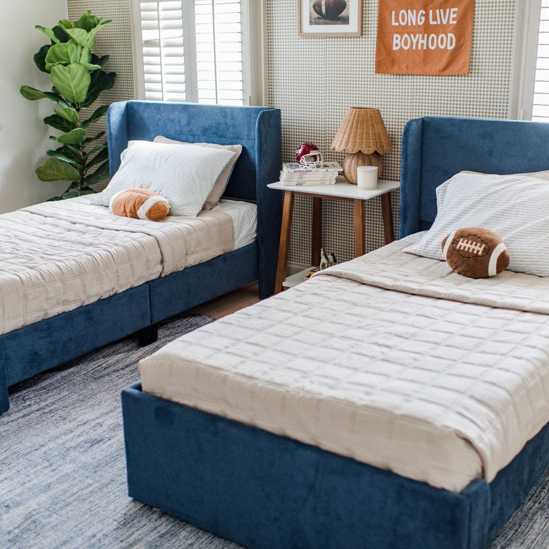 Twin beds with blue upholstered frames, football-themed pillows, and a minimalist nightstand in a stylish kid's bedroom.
