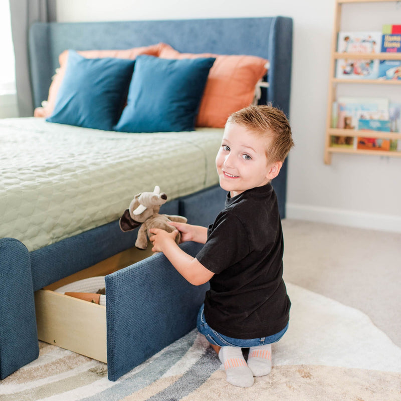Young boy organizing stuffed animals in a blue storage bed drawer in a bright, tidy bedroom.