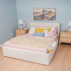 Cozy pastel bedroom with upholstered cream bed, pink bedding and patterned throw, mustard and blue accent pillows, plush bunny toys, wooden nightstands, matching lamps and abstract wall art