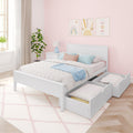 White wooden storage bed with two under-bed drawers in pastel pink children's bedroom with sunburst wall art, blue throw, bedside lamp and potted plant.