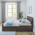 Bright modern bedroom with wooden platform bed and under-bed storage drawers, white bedding, pastel blue and lilac pillows, light blue throw, large window with sheer curtains, potted plant and textured blue wallpaper