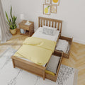 Wooden twin bed with pull-out under-bed storage drawers, yellow duvet and polka-dot pillow in a minimalist bedroom with nightstand, lamp, potted plant and framed fruit prints