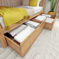 Under-bed storage drawers made of wood filled with folded linens and bedding, a space-saving bedroom storage solution next to a bed with a yellow throw and potted plant