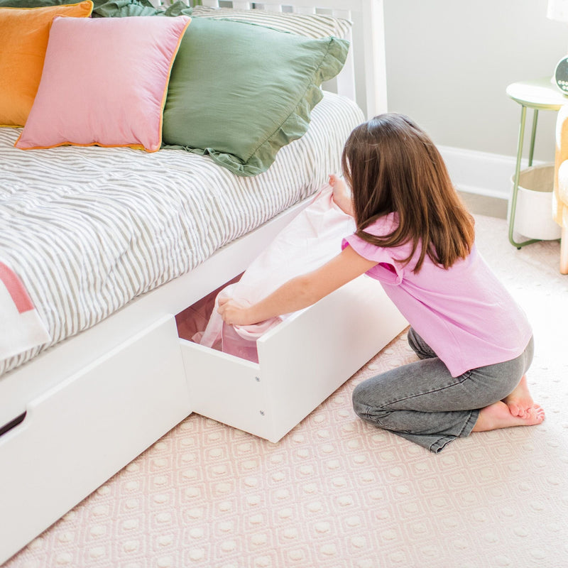 Young girl placing pink bedding into a white under-bed storage drawer in a bright kids' bedroom with striped sheets and colorful pillows.