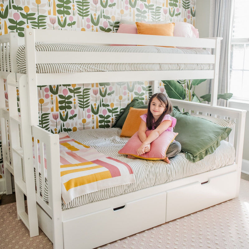 Cozy children's bedroom with white bunk bed and under-bed drawers, striped bedding, colorful floral wallpaper and throw pillows, child sitting on lower bunk holding a pink cushion by a sunlit window