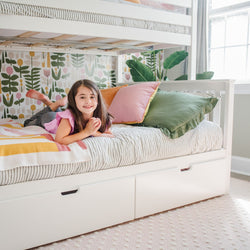 Girl smiling on a cozy white bunk bed with striped bedding and colorful pillows in a bright children's bedroom.
