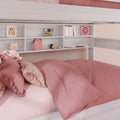 Blush pink bunk bed with rumpled duvet and ruffled pillows, white built-in shelves displaying books, a pink alarm clock, retro instant camera and portable speaker
