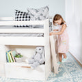 Young girl climbing ladder to white wooden kids loft bed with built-in storage, plush bunny, books and gray pillows.