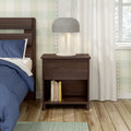 Dark walnut nightstand with gray table lamp and lower shelf of books next to bed with blue bedding on hardwood floor in modern bedroom interior