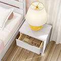 White nightstand with open drawer holding books and burlap pouch, yellow table lamp on top beside bed on wooden floor, modern cozy bedroom decor