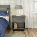 Grey wooden nightstand with modern dome lamp beside bed with blue bedding, books on lower shelf, white curtains, plaid wallpaper and hardwood floor