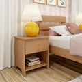 Cozy modern bedroom scene with a light wood nightstand (drawer and open shelf), yellow round table lamp, pink pillows and bedding on a wooden bed frame, plaid wallpaper and hardwood floor