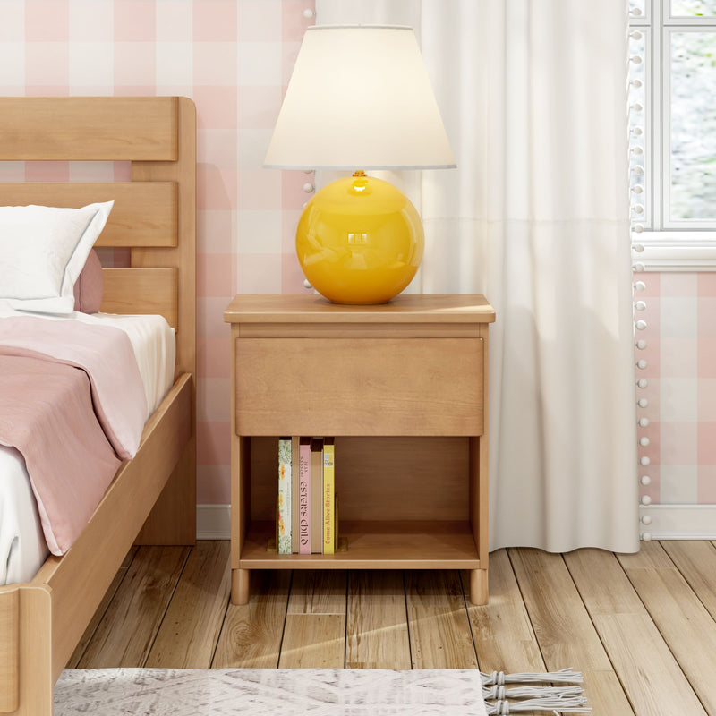 Wooden bedside table with yellow ceramic lamp next to light wood bed with pink bedding, pink gingham wallpaper, white curtains, books and hardwood floor