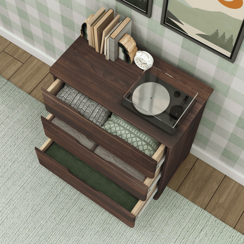 Top-down view of a mid-century modern dark wood dresser with three open drawers holding folded blankets and linens, a vinyl turntable, books and alarm clock on top, green plaid wallpaper and light green rug in a cozy bedroom setting