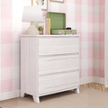 Whitewashed 3-drawer dresser in pink gingham nursery with beige table lamp, children's books and a green portable record player on top