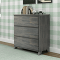 Gray three-drawer wooden dresser with tapered legs against mint-green gingham wallpaper, styled with books, alarm clock and vinyl player on a light wood floor and pale green rug