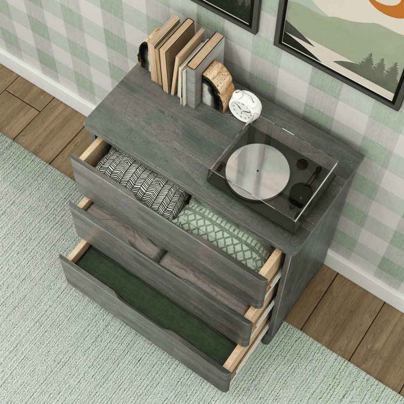 Gray wooden three-drawer dresser with open drawers showing folded blankets and linens, vintage turntable and books on top, white alarm clock, green plaid wallpaper, hardwood floor and green area rug — cozy bedroom storage and organization