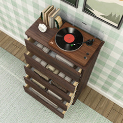 Mid-century modern walnut dresser with turntable record player and black vinyl, books and alarm clock on top, open drawers of folded blankets against green plaid wallpaper and hardwood floor