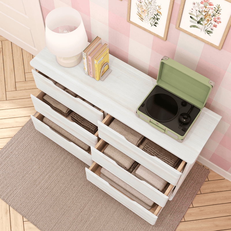 White wooden dresser with open drawers of folded blankets and linens, green portable record player on top, lamp, books and framed floral prints on a pink gingham wall — cozy bedroom/nursery storage and decor