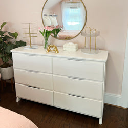 White dresser with six drawers, pink tulips in vase, modern decor, and round mirror reflecting a bedroom window.