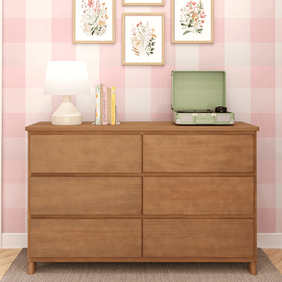 Wooden six-drawer dresser in pink gingham nursery with white table lamp, stacked books, mint green portable record player and framed floral wall art
