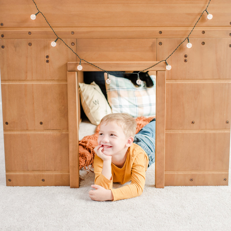 Smiling boy in a wooden indoor playhouse fort with plaid pillows and hanging globe string lights, cozy kids' play area