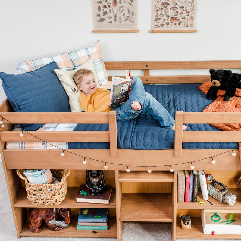 Child reading a book on a wooden loft bed with navy bedding and decorative pillows.