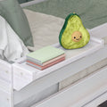 Plush smiling avocado pillow on a white wooden bed tray beside stacked pastel notebooks in a neutral kids’ bedroom