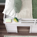 Top-down view of a white wooden kids' loft bed with safety rail, green pillow, white ruffled pillow, beige chevron blanket, avocado plush toy and stacked books on a bedside ledge over dark hardwood floor