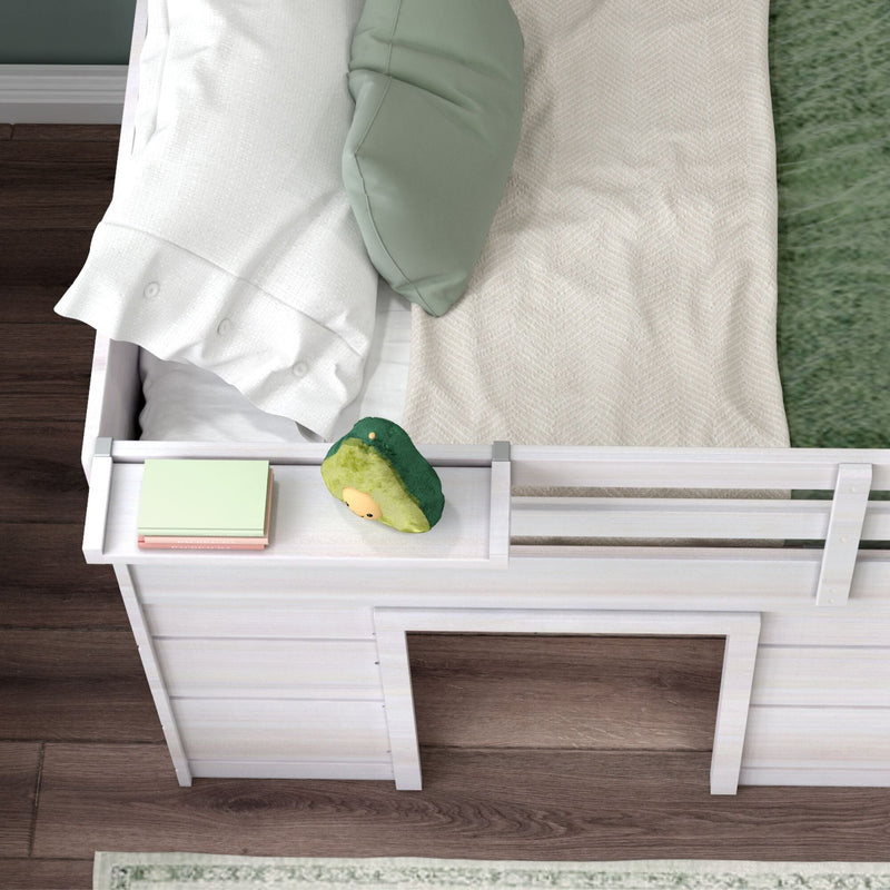 Top-down view of a white wooden kids' loft bed with safety rail, green pillow, white ruffled pillow, beige chevron blanket, avocado plush toy and stacked books on a bedside ledge over dark hardwood floor