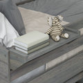 Gray wooden toddler bed with beige knit blanket, stack of books on the bed rail and striped elephant plush toy — neutral nursery decor