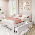 White wooden platform bed with pull-out trundle and blush pink bedding in a bright girls' bedroom with floral wall art, white curtains, bedside lamp, and matching dresser