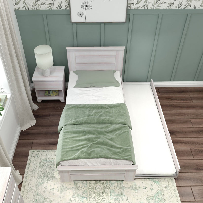 Whitewashed twin trundle bed with sage green bedding in farmhouse bedroom, paneled wall, nightstand lamp and hardwood floors