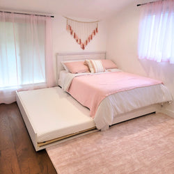 Pastel pink girls' bedroom with white wooden bed and pull-out trundle mattress, pink quilted bedding, decorative macramé wall hanging, sheer curtains, hardwood floors and blush area rug