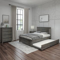 Modern farmhouse bedroom with grey wooden bed and pull-out trundle, matching dresser, neutral bedding, linen curtains framing a black-framed window, floral wallpaper accent, bedside lamp and dark hardwood floors