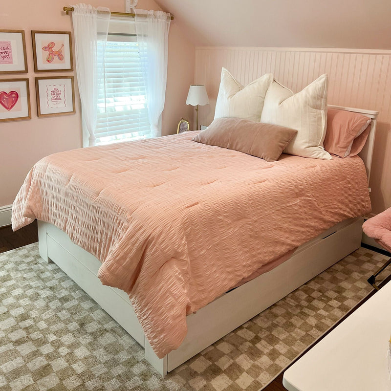 Blush pink bedroom with quilted pink comforter, white decorative pillows on a white platform storage bed, sheer curtains, framed wall art, beadboard wainscoting and beige checkered rug