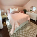 Cozy blush-pink bedroom with quilted bedspread, white decorative pillows, wooden nightstand, round dresser mirror, fresh tulips and potted plant
