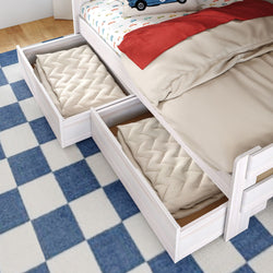 White wooden kids bed with two under-bed storage drawers pulled out, folded beige quilts inside, red throw and car-print sheet on mattress over blue checkered rug