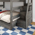 Grey wooden bunk bed with built-in staircase storage, beige bedding, car-print pillow and blue checkered rug in a kids' bedroom