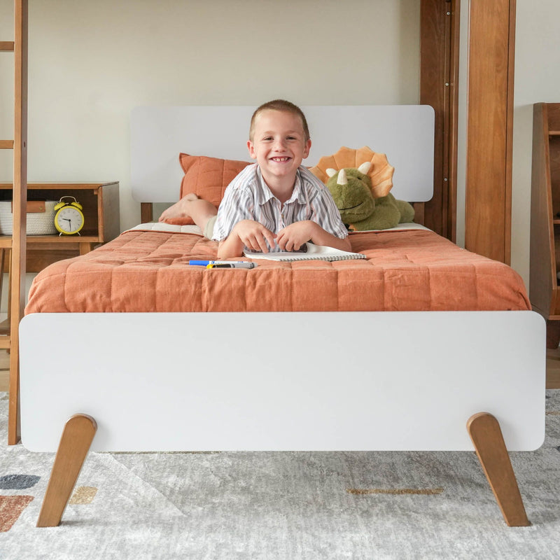 Child smiling while lying on a bed with an orange quilt, next to a stuffed dinosaur, in a modern bedroom setting.